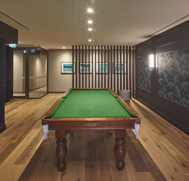 A refined billiards room with a full-size green-felt pool table centered on a wide timber floor. The room features dark painted wall panelling, a large botanical-patterned wall mural or artwork, framed artwork, glass doors, vertical timber screening, and recessed ceiling spotlights. The palette is warm timber, charcoal, and muted green, creating an upscale recreational interior.