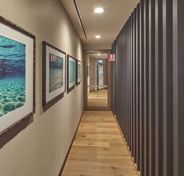 A narrow, warmly lit interior corridor with a light timber plank floor, textured neutral walls, framed ocean-themed artwork along the left side, and a tall dark vertical-slat screen forming the right wall. The hallway leads toward glazed doors and a brighter space beyond.