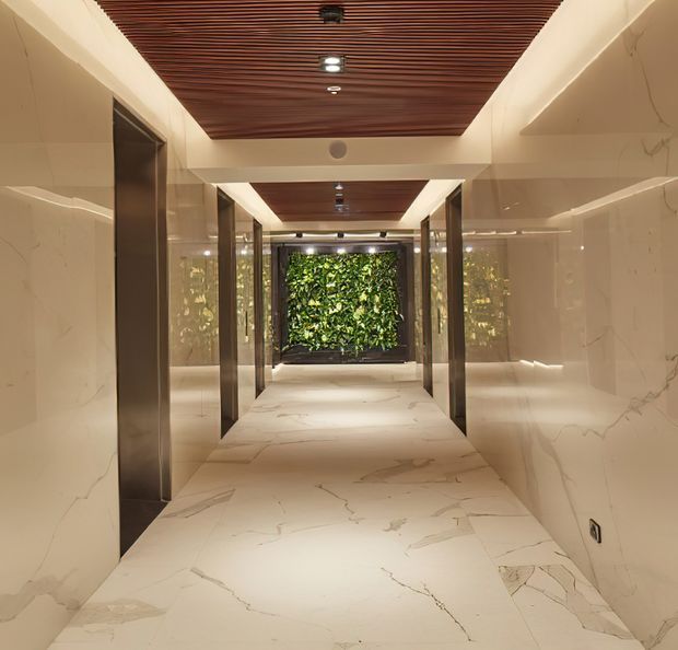 A luxurious contemporary interior corridor with large-format white marble-clad walls and flooring, dark metal-framed doorways, and a warm slatted timber ceiling with recessed lighting. The hallway leads toward a lush vertical garden feature, creating a refined, softly illuminated transition space.