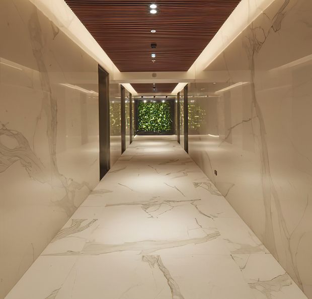 A refined, symmetrical interior corridor clad in large-format pale marble-effect surfaces on the walls and floor. Dark timber slatted ceiling panels with recessed lighting create contrast, while glazed openings and a planted green wall terminate the view. The hallway has a luxurious, contemporary character with clean lines and warm ambient lighting.