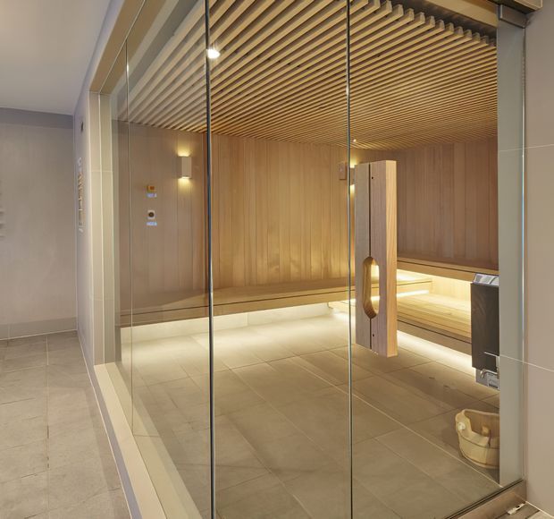 A refined sauna or steam room viewed through full-height clear glass doors. The space features warm timber wall panelling, a slatted timber ceiling, built-in tiered bench seating with concealed warm lighting, and large-format neutral floor tiles. A timber-framed glass door with an oval handle provides entry.