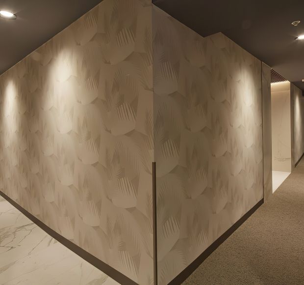A dimly lit contemporary interior corridor featuring a sculptural corner wall finished in pale textured patterned panels. The wall is illuminated by recessed spotlights and bordered by dark trim, with a pale marble-like floor on one side and carpeted flooring extending down the hallway. An open doorway and slatted ceiling detail are visible in the background.