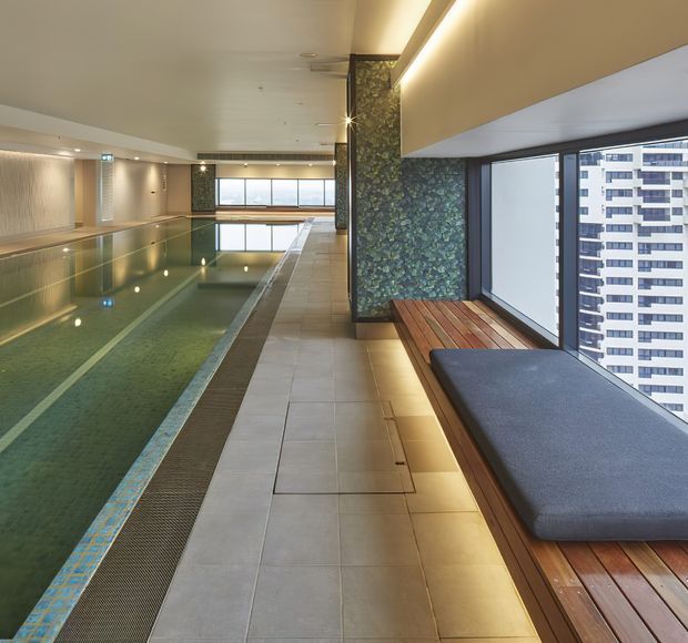 A sophisticated indoor lap pool in a high-rise building, with pale tiled flooring, full-height windows overlooking neighbouring buildings, integrated warm strip lighting, and a built-in timber bench with dark upholstered cushions. A green textured feature wall adds a natural accent beside the pool.