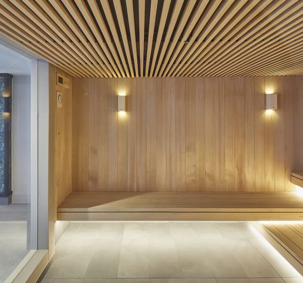 Warm contemporary timber-lined sauna interior with vertical wood wall panelling, a slatted timber ceiling, built-in tiered bench seating, soft wall lighting, and pale stone or tile flooring. A glazed opening is visible at the left.