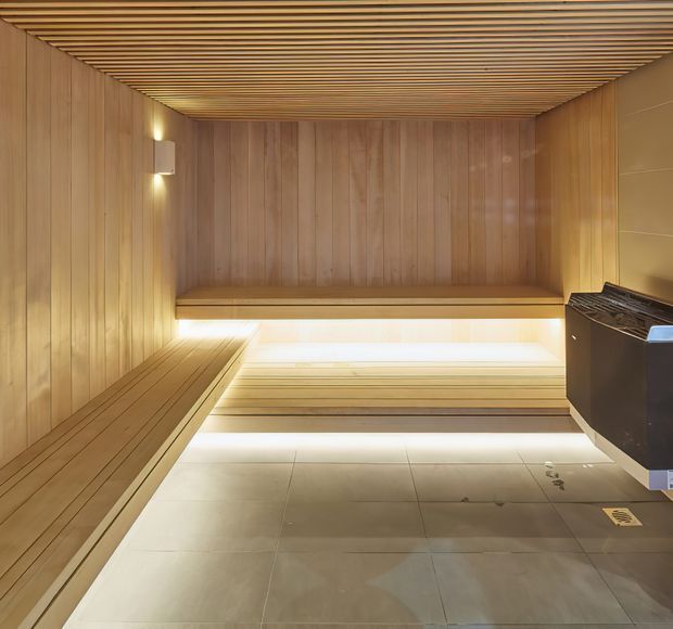 A compact modern sauna clad in warm timber panelling, with horizontal timber ceiling slats, built-in tiered benches, concealed linear lighting, and a dark sauna heater set against a tiled floor.