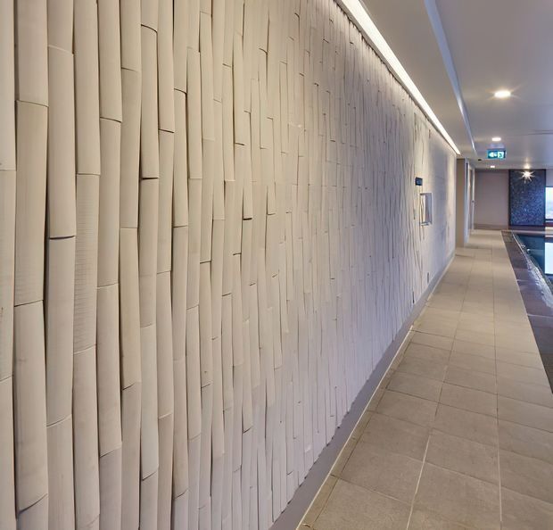 A long, softly lit interior corridor with a highly textured white sculptural wall made of narrow overlapping vertical panels. The corridor has pale tiled flooring, a smooth white ceiling with recessed spotlights, and a narrow water feature or pool visible along the right side. An illuminated exit sign and a dark patterned feature wall appear at the far end.