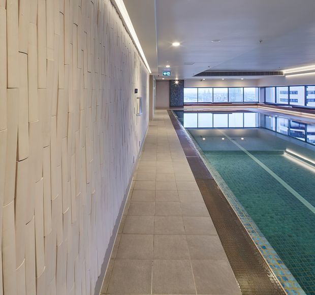 Interior view of a modern indoor swimming pool facility, showing a long tiled pool deck, turquoise tiled lap pool, large windows, recessed lighting, and a distinctive pale textured feature wall with vertical paneling.