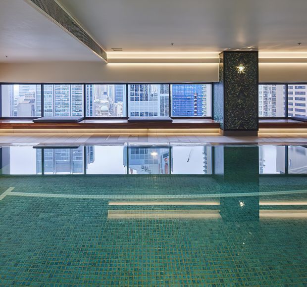 A modern indoor swimming pool with turquoise mosaic tile lining, reflecting the illuminated ceiling and large city-facing windows. A dark patterned feature column divides the glazing, with built-in timber-toned bench seating along the perimeter. Dense high-rise buildings are visible outside, creating an urban wellness or recreation setting.