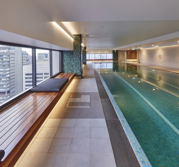 A sleek indoor lap pool in a high-rise building, with turquoise tiled water, marked swimming lanes, a tiled pool deck, timber bench seating along floor-to-ceiling windows, and a textured feature wall. City apartment or office towers are visible through the glazing.