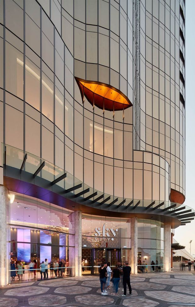 Exterior view of a contemporary multi-storey commercial building with a curved, highly glazed facade and distinctive projecting amber-orange canopy. The ground level contains a brightly lit hotel or entertainment entrance with visible signage, cylindrical stone-clad columns, a deep glass canopy, and patterned paved forecourt. People are present near the entry.