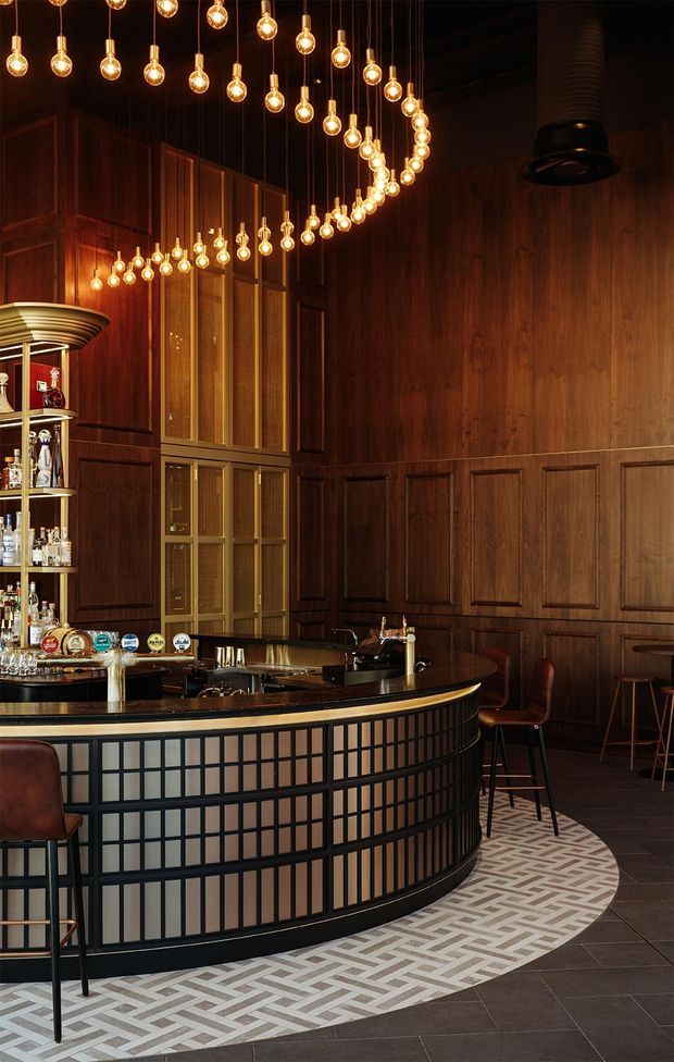 A sophisticated dark-toned cocktail bar with rich timber wall panelling, a curved black-framed bar front with pale inset panels, brass detailing, integrated bottle shelving, and a dramatic canopy of hanging globe pendant lights. A patterned geometric tile border defines the bar area against the dark floor, while bar stools and a large black ventilation hood complete the commercial hospitality setting.