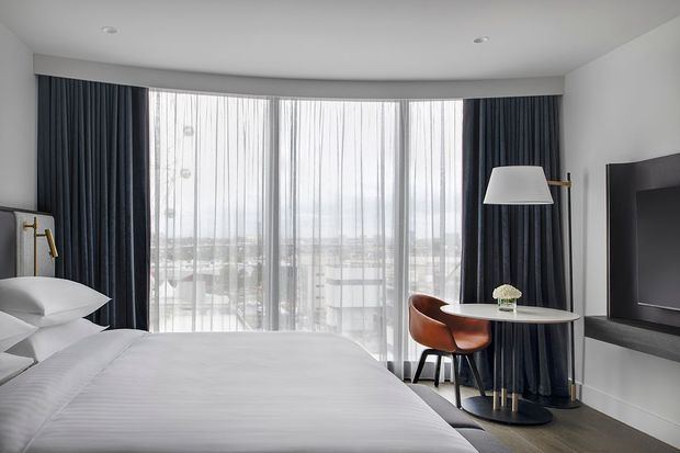 A refined modern bedroom with a curved floor-to-ceiling window wall framed by dark navy curtains and sheer voile drapes. The room features a neatly made white bed, a small round table with a brown leather chair, a minimalist floor lamp, wall-mounted television, pale built-in cabinetry, and dark timber-look flooring. The outlook through the glazing is visible but secondary.