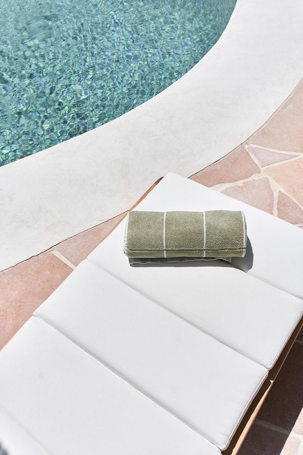 A close-up of a white cushioned poolside sun lounger with a folded olive-green striped towel, beside a curved swimming pool edge and pale terracotta stone paving. The image is bright, minimal, and resort-like.