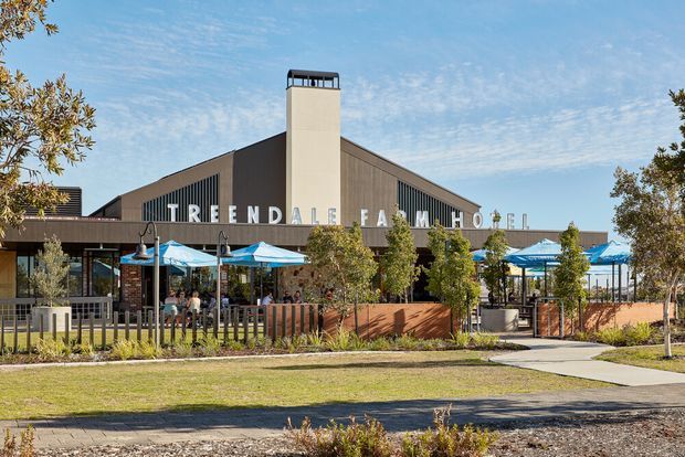 Exterior view of a contemporary gabled commercial venue with a tall central chimney or tower, dark vertical cladding, and large lettering reading “Treendale Farm Hotel.” The building fronts an outdoor hospitality area with blue umbrellas, seating, timber fencing, landscaped planting, and a pedestrian path.