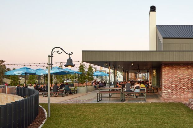 Exterior view of a contemporary commercial hospitality venue with a covered outdoor dining terrace. The building combines a dark standing-seam metal roof and canopy, exposed red brick walls, large glazed openings, and pale vertical cladding. The terrace contains picnic-style tables, umbrellas, decorative street lamps, and landscaped lawn areas, with additional seating visible inside.
