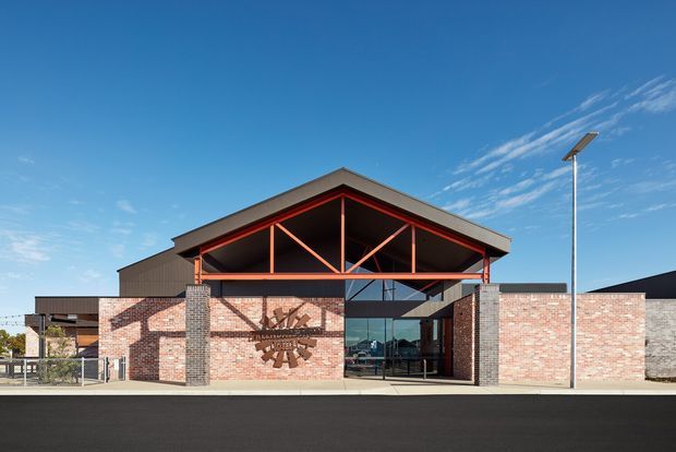Front exterior view of a low-rise contemporary commercial building with extensive exposed red brick walls, dark masonry columns, and a prominent open gable entrance canopy supported by exposed steel trusses painted orange-red. Glazed entry doors sit beneath the canopy, with a paved forecourt and tall solar streetlight visible against a clear blue sky.