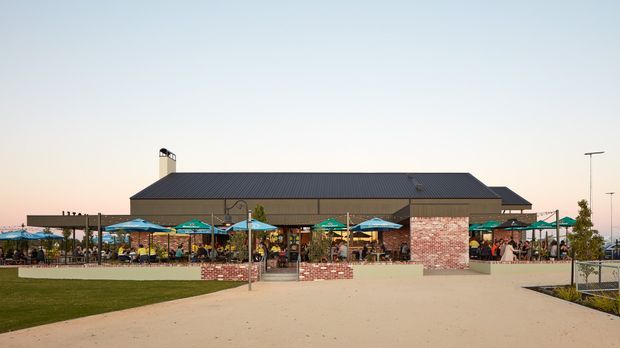 Wide exterior view of a low-rise commercial venue with a large dark pitched roof, broad covered outdoor dining area, colorful umbrellas, brick and pale rendered wall sections, paved forecourt, and landscaped lawn. The building appears to function as a hospitality or retail space, with numerous patrons seated outdoors.