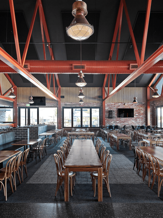 A spacious industrial-style restaurant or dining hall with long wooden communal tables, numerous bentwood chairs, dark carpet flooring, exposed red-painted steel roof trusses and ductwork, black ceiling panels, pendant lights, exposed brick walls, and large glazed doors and windows. A bar or booth seating area and wall-mounted screens/fireplace-like features are visible in the background.