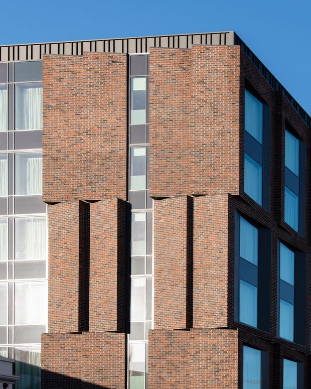 Close-up exterior view of a multi-storey building with a strongly articulated red-brown brick facade. Projecting and recessed brick volumes create a rhythmic geometric composition around narrow vertical strips of glazing, while larger blue-tinted windows appear on the darker side elevation. A black metal parapet or railing runs along the flat roofline against a clear blue sky.