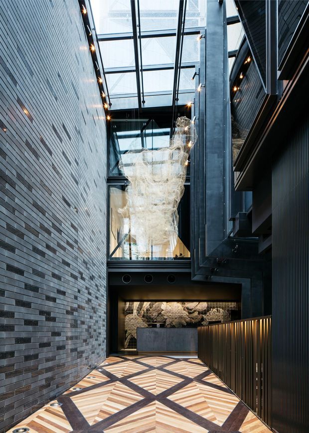 A tall, dramatic interior atrium or entry passage with a glazed roof and multi-level circulation. Dark linear brick walls contrast with black metal structural framing and ribbed wall panels. A large translucent sculptural pendant hangs through the void, while a patterned timber floor forms geometric diamond motifs. Warm integrated lighting highlights the artwork and walkway, creating a refined, contemporary commercial or hospitality atmosphere.