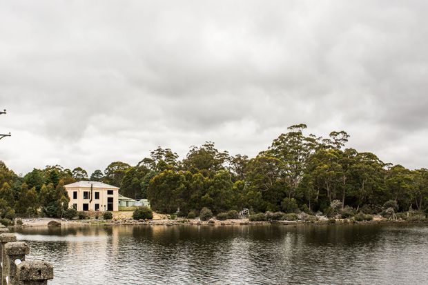 A distant exterior view of a pale multi-storey house beside a calm body of water, surrounded by dense mature trees and rocky shoreline under an overcast sky. The image is primarily a landscape and waterfront setting rather than a detailed view of the building.