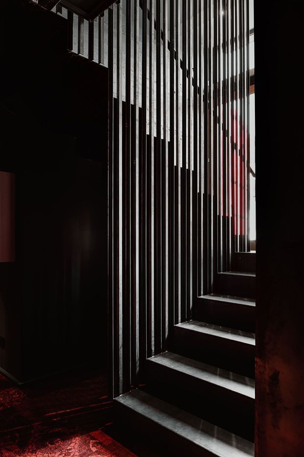 Dramatic, moody interior photograph of a dark stairway with black steps and a striking vertical slatted screen or balustrade. Narrow shafts of daylight enter from the right, creating strong linear shadows and highlighting the textured dark surfaces. A red patterned floor covering and warm red reflections add a subtle accent.