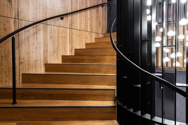 Interior architectural photograph of a contemporary stairway with broad warm timber treads and matching timber wall panelling. Slim black metal handrails curve up the stair, while a black steel and glass balustrade opens to a darker adjacent space with multiple hanging pendant lights.