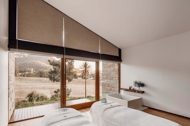 A serene, minimalist spa-like bedroom with two beds positioned beside full-height glazing. The room features white bedding, a freestanding white bathtub, pale plaster walls, dark timber window framing, and a stone masonry wall section. Exterior landscape is visible through the glazing.