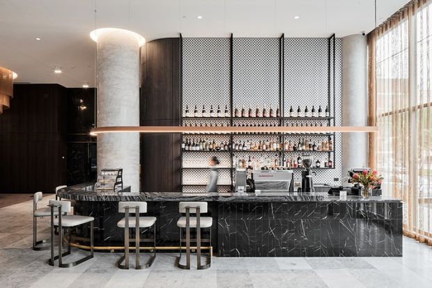 A refined contemporary bar and reception-style counter with a long black veined stone island, pale upholstered stools, dark timber wall panelling, a cylindrical textured column, and an open display of bottles against patterned screens. Floor-to-ceiling windows with sheer curtains line the right side, bringing in abundant natural light.