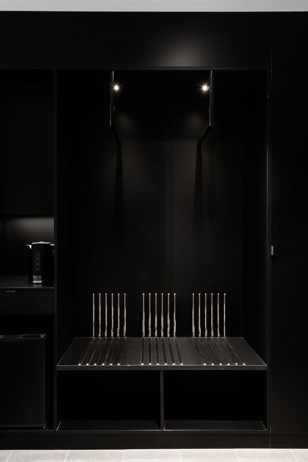 A dramatic, highly concealed black built-in joinery installation, likely an entryway or wardrobe area, with a stainless-steel slatted bench and matching vertical hooks beneath two small spotlights. Dark cabinetry and wall panels create a sleek, minimalist composition. A black electric kettle and compact appliance are visible in an adjacent open compartment at left. Pale tiled flooring provides the only strong contrast.