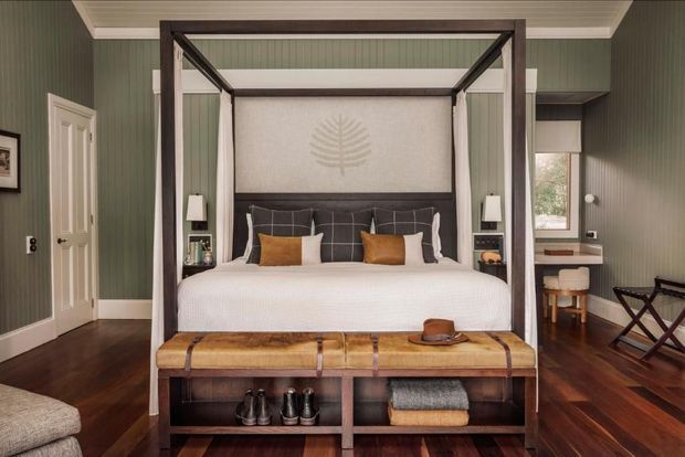 A refined bedroom with a dark timber four-poster canopy bed, upholstered grey headboard, crisp white bedding, and warm tan accent cushions. The room features green vertical wall panelling, a white beadboard ceiling, dark timber flooring, built-in bedside niches, and a substantial timber storage bench at the foot of the bed. A small desk and folding chairs sit beside the window.