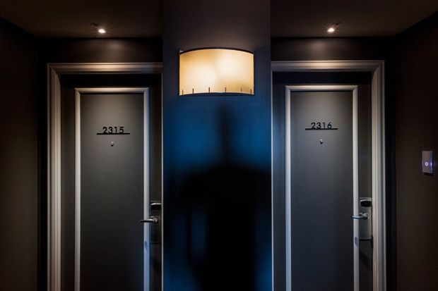 A dimly lit hotel or apartment corridor showing two dark grey entrance doors numbered 2315 and 2316. The doors have metallic trim and handles, recessed ceiling spotlights, and a central dark feature wall with a softly glowing wall light. A small illuminated access panel is visible on the right.