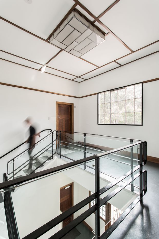 A bright multi-level interior landing or stair hall with a glass balustrade and black metal handrails surrounding an open stairwell. White walls and ceiling panels are outlined with dark timber trim, complemented by timber doors, a large gridded window, and a geometric suspended ceiling light. The space has a refined modern-industrial character and a strong double-height void.