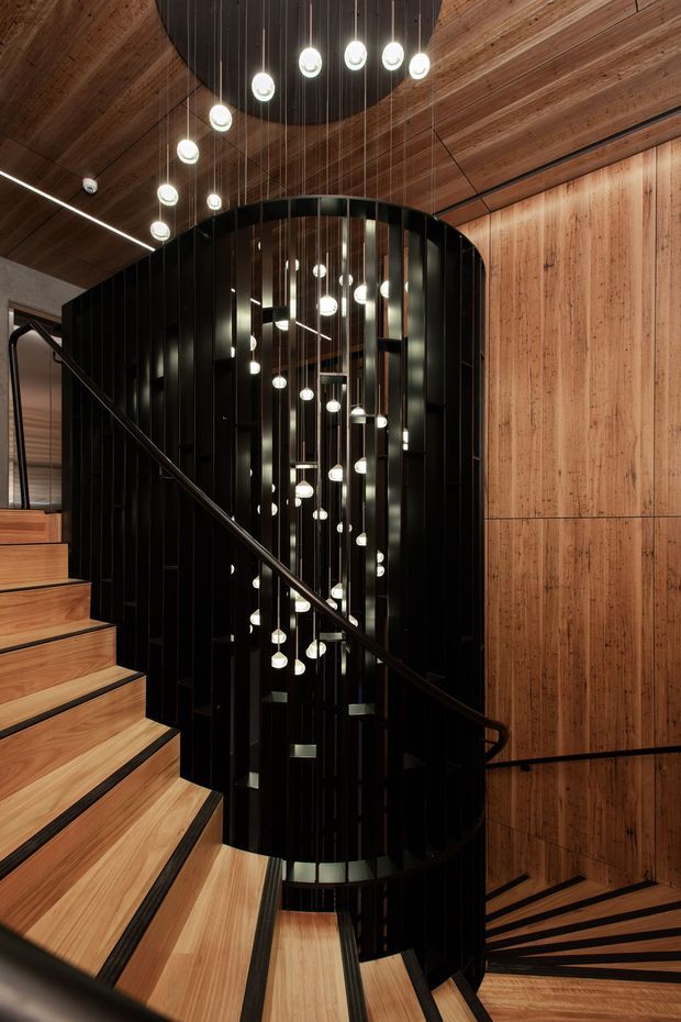 Dramatic contemporary stairway with curved black metal screening and a sculptural cascade of spherical pendant lights. Warm timber cladding covers the walls and ceiling, while timber stair treads are edged with dark contrasting strips. A black handrail follows the sweeping stair geometry, and reflective surfaces amplify the lighting within the stair void.
