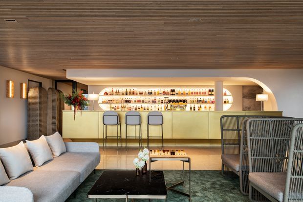 A sophisticated contemporary home bar and lounge with a long backlit bottle display, pale green bar cabinetry, three stools, and an integrated curved alcove. The foreground contains a large light-grey sectional sofa, black coffee table, and woven lounge chairs. A richly textured timber-lined ceiling, polished stone-like flooring, soft ambient lighting, and dark green rug create a warm, refined hospitality-inspired atmosphere.