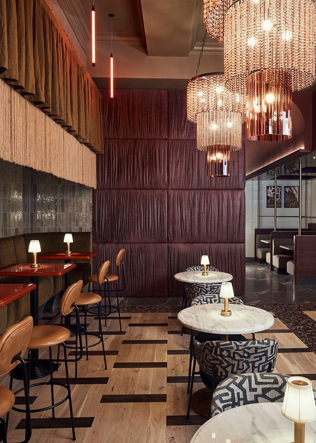 A sophisticated hospitality interior, likely a restaurant or hotel lounge, with richly layered materials and dramatic lighting. The space features burgundy padded wall panels and drapery, dark glossy tiled walls, patterned timber flooring, marble café tables, dark upholstered banquettes, tan leather bar stools, and several large cascading crystal chandeliers. A glazed partition reveals additional booth seating in the background. The atmosphere is opulent, moody, and refined, with strong Art Deco-inspired detailing.