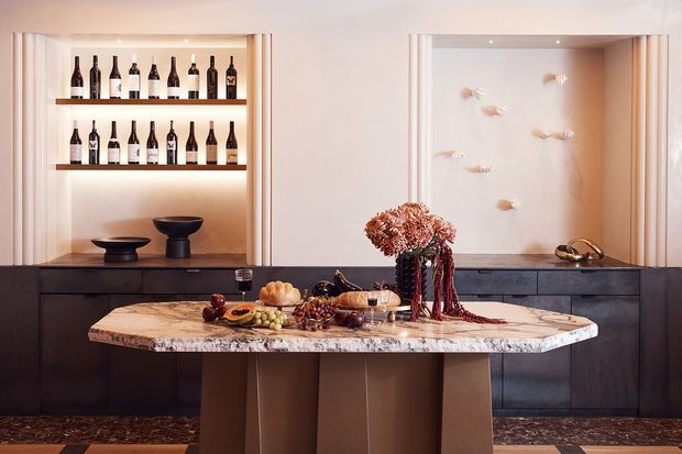 A sophisticated dining and entertaining area with a large marble-topped island or buffet table in the foreground. Dark built-in cabinetry spans the rear wall, incorporating illuminated wine bottle shelving on the left and a recessed decorative display niche on the right. Sculptural vessels, floral arrangements, tableware and food create a refined, styled setting, with warm integrated lighting and contrasting dark cabinetry.