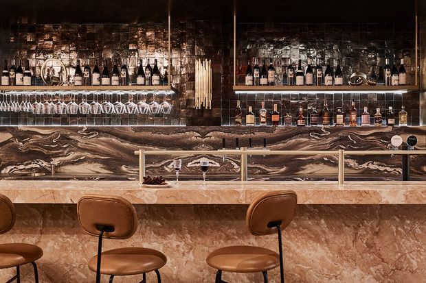 A sophisticated bar interior with a long marble-clad counter, sculptural veined stone backsplash, brass or dark metal trim, illuminated open shelving displaying bottles and glassware, and several brown leather bar stools. The space has a dramatic, luxurious, richly layered atmosphere with warm low lighting and reflective surfaces.