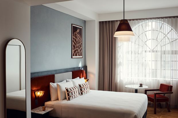 A refined bedroom with a large upholstered bed, white bedding, patterned accent cushions, and a dark timber headboard set against a textured grey feature wall. Warm bedside lighting, an arched full-length mirror, sculptural wall art, floor-length curtains, and a large arched window create an elegant, softly lit interior.