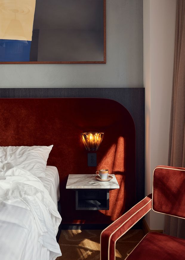 A richly styled bedroom corner featuring a bed with rumpled white bedding, a dramatic deep burgundy upholstered wall and integrated rounded headboard, a wall-mounted amber glass sconce, and a small marble bedside table with open storage. A matching burgundy velvet armchair sits beside tall curtains, while warm sunlight creates strong highlights and shadows. The scene has a boutique-hotel, refined and moody character.