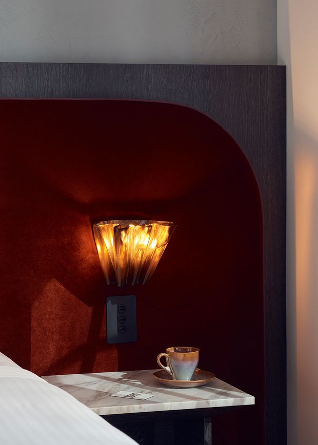 Close-up of a richly styled bedroom bedside alcove. A dark timber-toned surround frames a deep burgundy curved inset wall with an illuminated sculptural wall sconce, beside a marble bedside table holding a cup and saucer. The edge of a white bed is visible in the foreground, creating a warm, intimate, luxurious atmosphere.