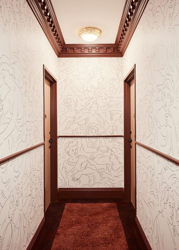 A narrow, symmetrical hallway with richly stained dark timber door frames, skirting, dado rails, and an ornate coffered ceiling trim. The walls feature pale wallpaper with line-drawn figurative artwork, while a deep red-brown carpet runner and vintage-style ceiling light create a dramatic, theatrical atmosphere.