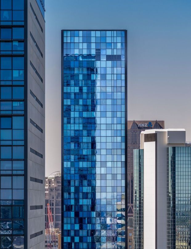Vertical cityscape photograph showing several tall commercial office buildings, dominated by a narrow rectangular tower with a blue reflective glass curtain-wall facade and regular square grid. Adjacent buildings feature dark blue glazing, pale concrete or metal panels, and modern geometric forms. The image is captured outdoors in clear daylight with an urban skyline context.