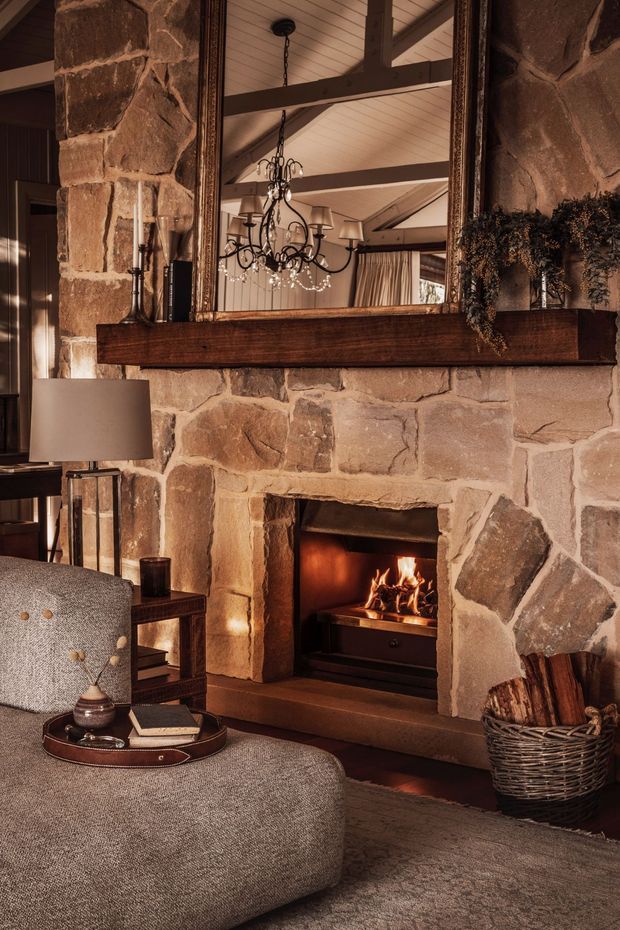 A warm, rustic living room centred on a substantial rough-hewn stone fireplace with a lit fire. A large framed mirror with a dark timber mantel reflects exposed timber ceiling beams, a traditional chandelier, and curtains. The room includes a textured upholstered lounge, side table, floor lamp, and woven firewood basket, creating a cosy lodge-like atmosphere.