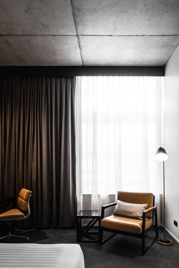 A moody, refined bedroom corner with a low concrete ceiling, dark full-height curtains, sheer window drapery, charcoal carpet, and minimalist black-framed timber seating. A floor lamp stands beside an armchair, while the edge of a bed is visible in the foreground. The space combines raw architectural surfaces with warm leather upholstery and dramatic contrast.
