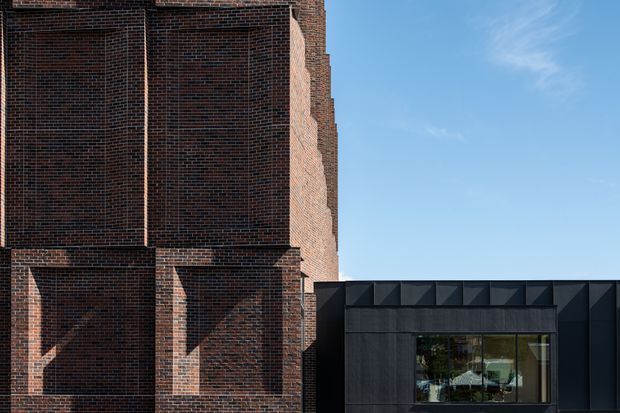 Exterior architectural photograph showing a dark red-brown brick building with strong projecting vertical and horizontal brick pilasters, joined to a lower contemporary black-clad volume with a large glazed window. Clear blue sky and crisp sunlight emphasize the contrast between the masonry structure and the modern addition.