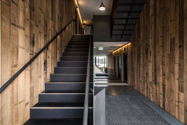 A narrow industrial-style interior corridor with a straight black staircase rising along the left wall and a dark metal mezzanine or overhead stair structure above. Both side walls are clad in rough reclaimed timber boards, contrasted with black metal stair treads, handrails, framing, and a textured dark floor. Warm linear lighting and pendant lights create a dramatic, moody atmosphere, with glazing visible at the far end.