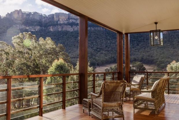 Covered timber-framed outdoor terrace with extensive glazing and open sides, furnished with woven wicker lounge chairs around a small table. The timber deck and warm wood ceiling overlook a dramatic forested mountain escarpment.