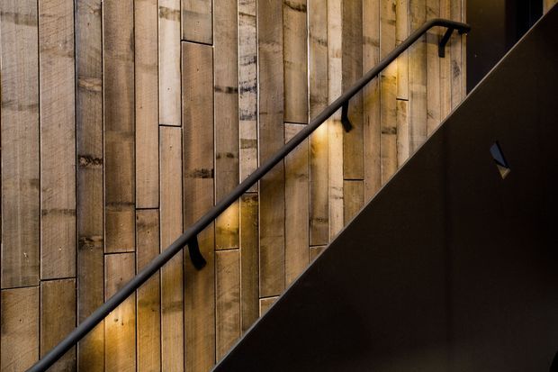 Interior stairway detail with a black metal handrail running diagonally alongside a dark stair balustrade. The wall is clad in vertically arranged reclaimed or rustic timber boards, with warm concealed lighting creating a dramatic, atmospheric effect.