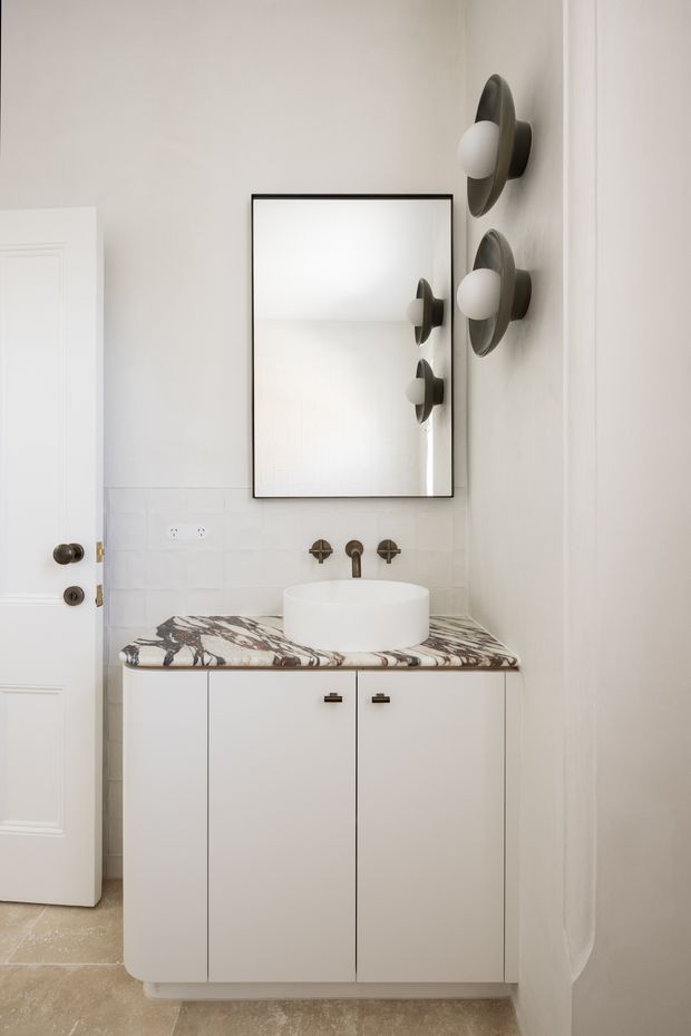 A compact, refined bathroom vanity area with a white vessel basin on a dramatic veined stone benchtop. The vanity has simple white flat-panel cabinetry, a wall-mounted aged-metal tap set, a large black-framed rectangular mirror, and sculptural dark wall sconces with round white globes. Light tiled walls and a pale stone-look floor create a bright, softly neutral interior.